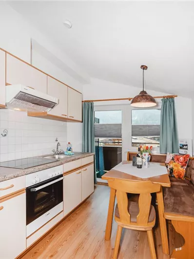 A modern kitchen with wooden cabinets and a dining area. Large windows provide plenty of daylight and a view outside.