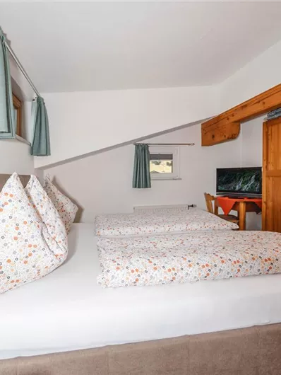 A cozy bedroom with two beds and bright pillows. On the wall, there is a wooden wardrobe and a television.