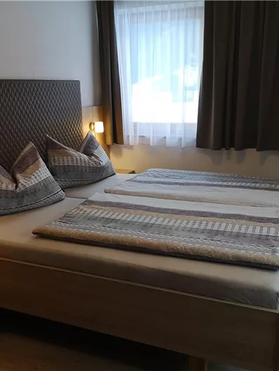 A cozy bedroom with a large double bed and two pillows. Bright curtains and a chair complement the decor.