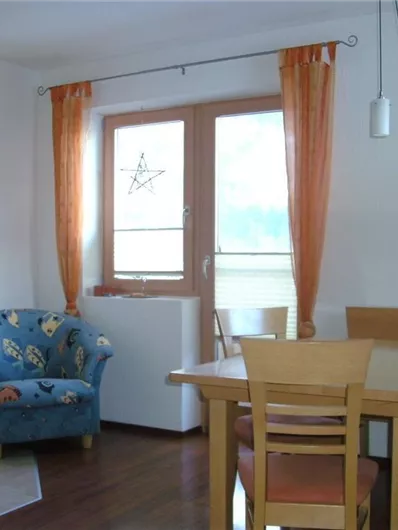A cozy dining area with a wooden table and chairs. In the background, there is a blue armchair and large windows with orange curtains.