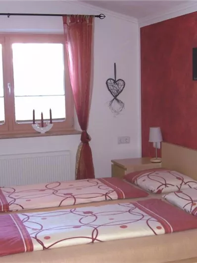 A cozy bedroom with two single beds and red curtains. A decorative picture hangs on the wall, and a small bedside table stands next to the beds.