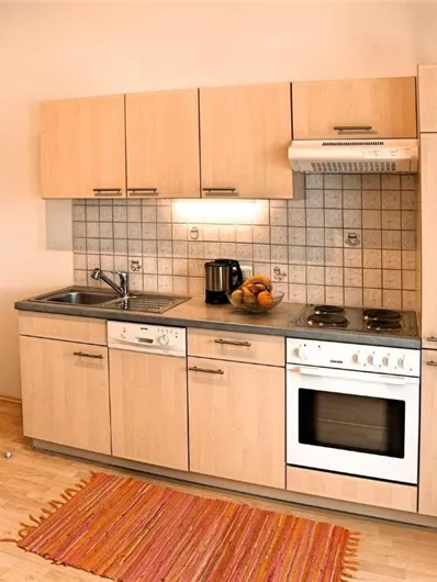 Een heldere keuken met moderne houten kasten en lichte muren. Op de vloer ligt een kleurrijk tapijt en op het aanrecht staan een koffiekan en enkele vruchten.