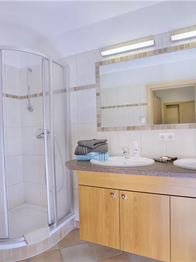 Een moderne badkamer met een douche en twee wastafels. De wanden zijn licht betegeld en er is een houten kast met handdoeken.