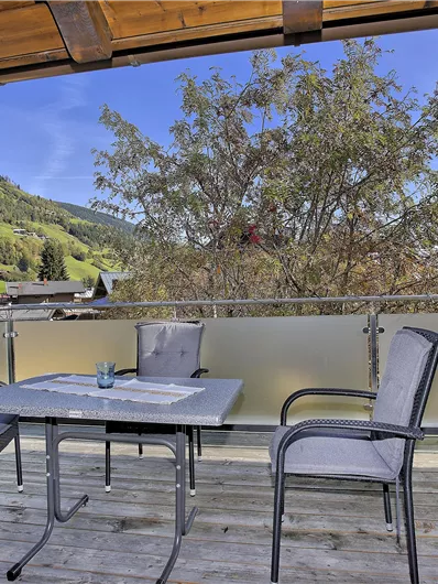 Een zonnig balkon met een tafel en twee stoelen. Op de achtergrond zijn groene bergen en een heldere blauwe lucht te zien.