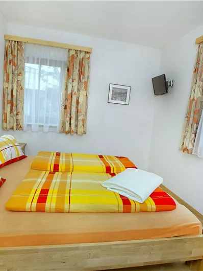 Een gezellige slaapkamer met een tweepersoonsbed en kleurrijk beddengoed. De kamer heeft grote ramen en een lichte, uitnodigende sfeer.