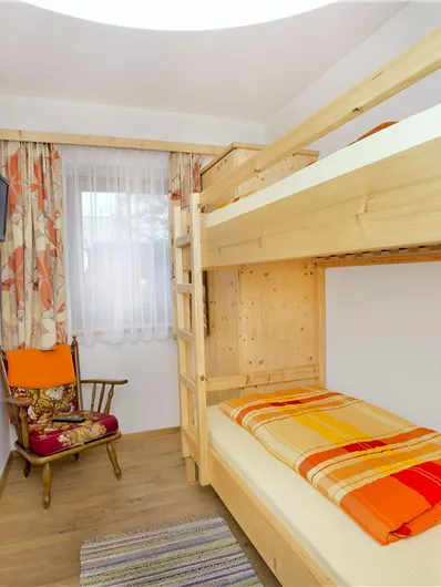 Een eenvoudige kamer met een hoogslaper en een fauteuil. Er is een raam met gordijnen en een televisie aan de muur.