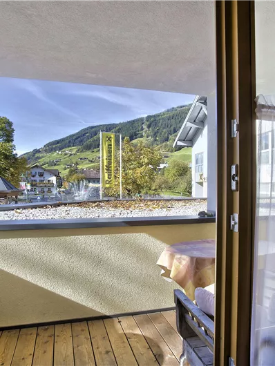 Een balkon met een tafel en twee stoelen. Op de achtergrond zijn groene bergen en een blauwe lucht te zien.