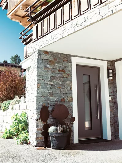 A modern entrance portal with a stylish door and a cozy outdoor area. The surroundings are filled with plants and a clear blue sky.