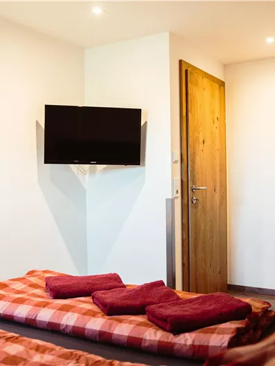 A cozy room with two beds, red bedding, and a television on the wall. In the background, there is a wooden door and a picture on the wall.