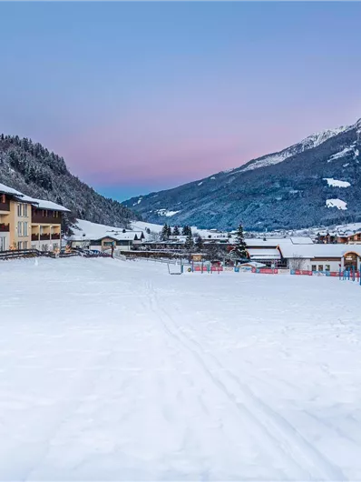 Eine verschneite Landschaft mit gemütlichen Chalets und majestätischen Bergen im Hintergrund. Der Himmel hat einen sanften Farbverlauf in der Dämmerung.