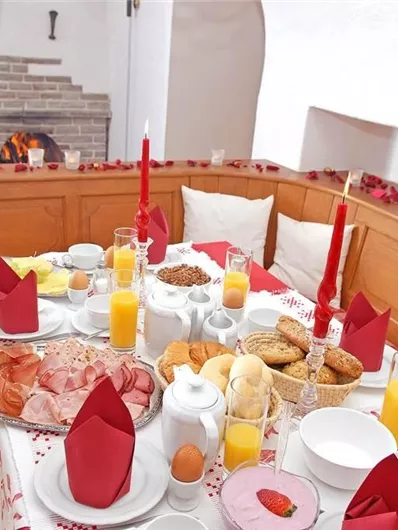 Ein festlich gedeckter Tisch mit rotem Tischdecke und dekorierten Kerzen. Die Speisen umfassen Aufschnitt, Brötchen, Kekse und Säfte, ideal für ein gemütliches Frühstück.