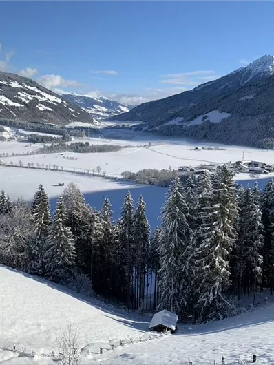 Eine schneebedeckte Landschaft mit hoch aufragenden Bergen und einem klaren blauen Himmel. Im Vordergrund sind schneebedeckte Bäume und eine Wiese zu sehen.