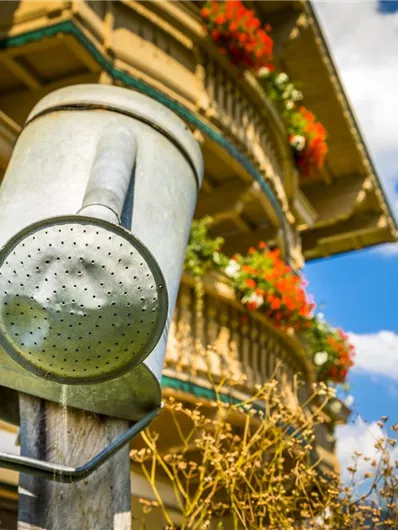 Een zilveren gietkan hangt aan een houten paal. Op de achtergrond ziet men een gebouw met bloemen en een blauwe lucht.
