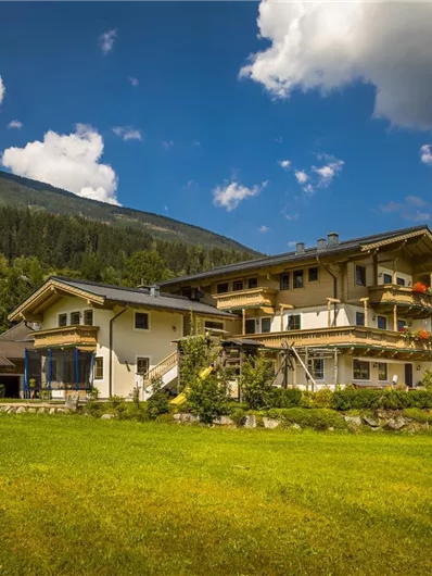 Een groot, modern huis omgeven door groene grasvelden en bomen. Op de achtergrond zijn zachte heuvels en een blauwe lucht met enkele wolken te zien.