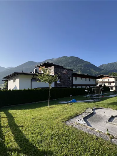A quiet landscape with several buildings and green meadows. In the background, majestic mountains rise under a bright blue sky.