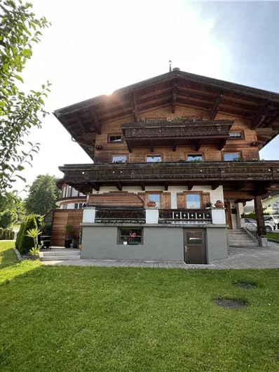 A traditional alpine house with wooden balconies and a well-maintained garden. The surroundings are green and surrounded by mountains.