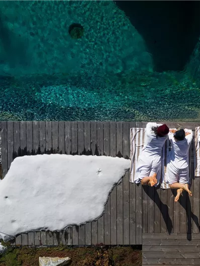 Three people are lying on a wooden terrace next to a pool. In the background, a bit of snow is still visible.