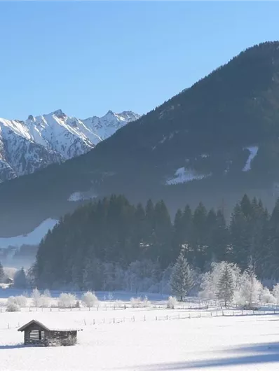 Een besneeuwd winterlandschap met hoge bergen op de achtergrond. Op de voorgrond staat een klein, zwart huisje en enkele besneeuwde bomen.