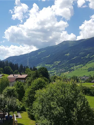 Eine malerische Landschaft mit grünen Hügeln und einem klaren Himmel. Im Vordergrund sind einige Häuser und Bäume sichtbar.