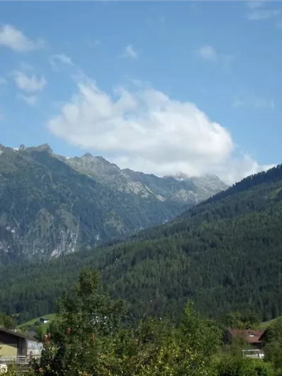 Een indrukwekkend berglandschap met groene bossen en gezellige huizen op de voorgrond. De lucht is blauw met enkele wolken.