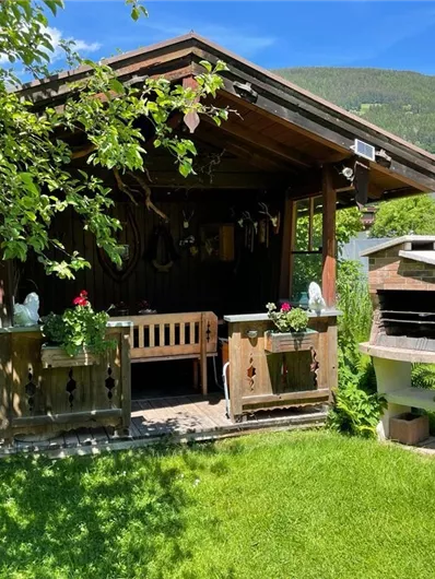 A cozy garden shed surrounded by green grass and various plants. Next to the shed is a barbecue area with a brick grill.