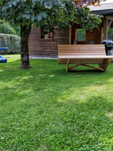 A well-kept garden with green grass and a wooden bench. In the background, there is a wooden house with a slide and a grill.