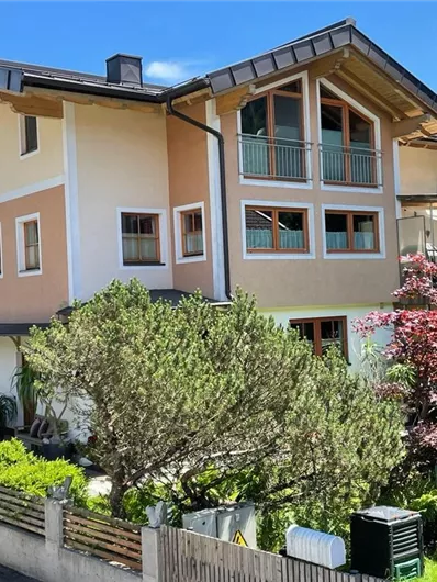 A modern house with a bright facade design and balconies. The garden is green and well-maintained, surrounded by beautiful plants.