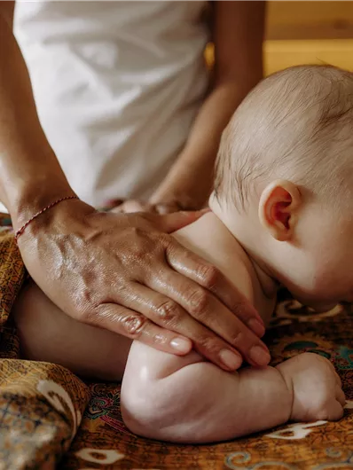 Ein Baby liegt auf einem Tisch und bekommt eine sanfte Massage. Eine Hand hilft dabei, das Baby zu entspannen.