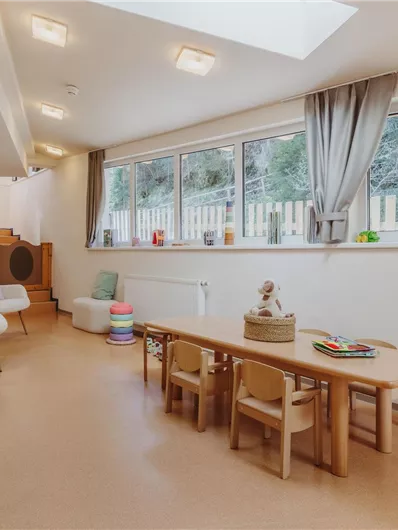 Ein helles Spielzimmer mit großen Fenstern und einem freundlichen Ambiente. Es gibt einen Tisch mit Stühlen und verschiedene Spielzeuge in der Nähe.