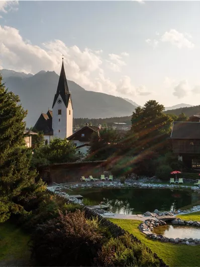 A picturesque landscape with a church and mountains in the background. The garden with a pond and loungers offers a relaxed atmosphere.