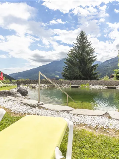 A beautiful outdoor pool in the midst of nature with green meadows and mountains in the background. Plastic loungers are ready, while some sunshades provide shade.
