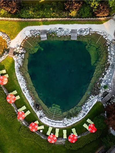 A beautiful, natural pool amidst green grass and trees. Around the pool, deck chairs and sun umbrellas are placed.