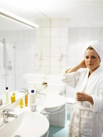 A woman in a bathrobe stands in the bathroom and looks into the mirror. Various cosmetic products are placed on the sink.