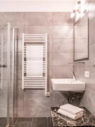Ein modernes Badezimmer mit einer Dusche und einem Waschtisch. Die Wände sind in grauen Fliesen gehalten und ein Handtuch liegt bereit.