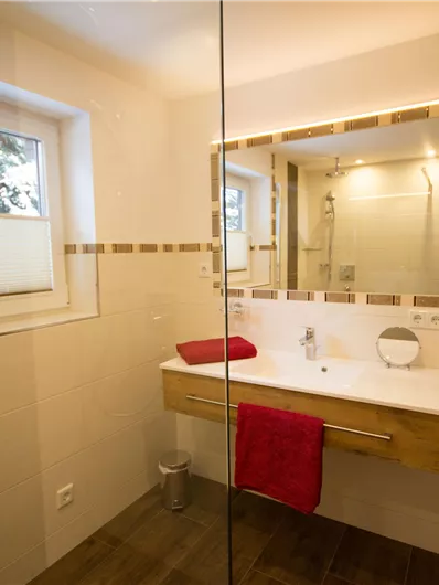 A modern bathroom with a spacious shower and a bright mirror. The room is tastefully decorated with wood and red accents.