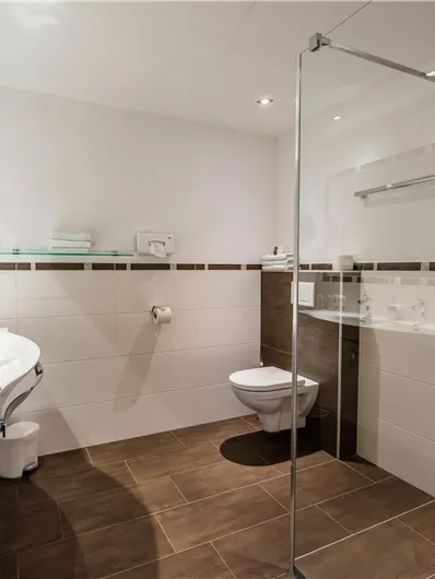Ein modernes Badezimmer mit einer Glasdusche und einem weißen Waschbecken. Die Wände sind hell gefliest und der Boden hat einen dunklen Farbton.