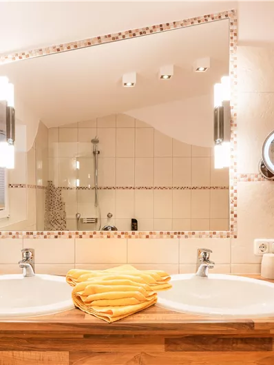 Ein modernes Badezimmer mit zwei Waschbecken und einem großen Spiegel. Handtücher in Gelb liegen ordentlich auf der Holzablage.