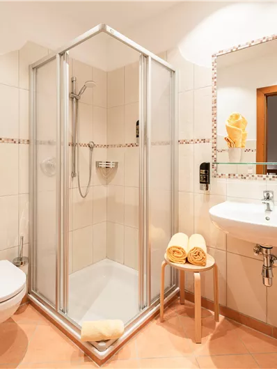 Ein modernes Badezimmer mit Dusche und Waschbecken. Die Wände sind hell gefliest, und es gibt eine kleine Ablage mit Handtüchern.