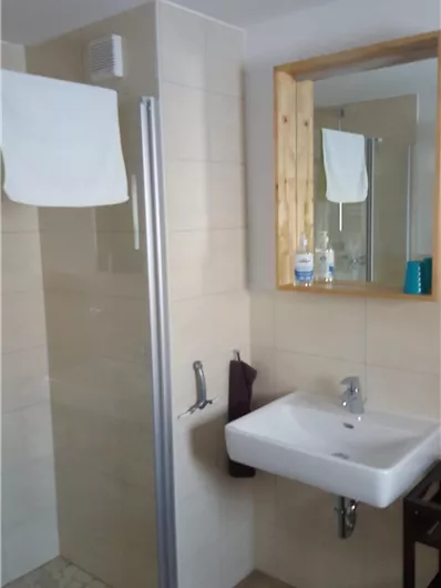 Ein modernes Badezimmer mit einer Duschkabine und einem Waschbecken. Im Hintergrund befindet sich ein Spiegel und Handtücher sind aufgehängt.