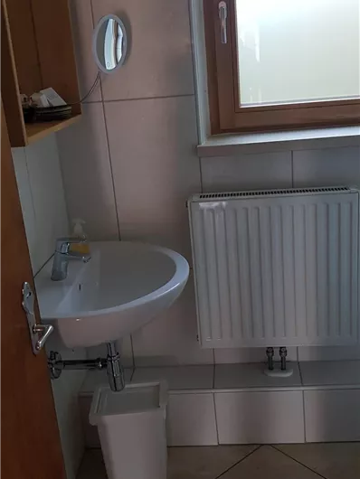 Ein kleines Badezimmer mit einem Waschbecken, einem Heizkörper und einem Fenster. Der Boden ist mit Fliesen ausgelegt und es gibt einen Abfalleimer.