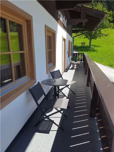 Ein gemütlicher Balkon mit einem kleinen Tisch und Stühlen. Im Hintergrund ist eine grüne Wiese und ein klarer Himmel zu sehen.