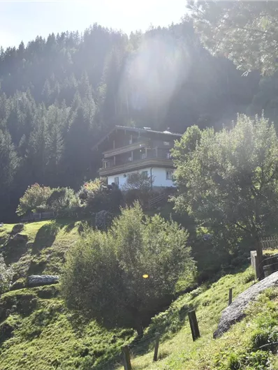 Ein malerisches Haus auf einem grünen Hügel, umgeben von Bäumen. Im Hintergrund erheben sich hohe Berge unter einem klaren Himmel.
