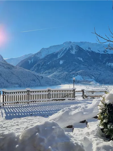 A snowy landscape with mountains in the background and radiant sunshine. In the foreground, there is a wooden terrace and snow-covered plants.