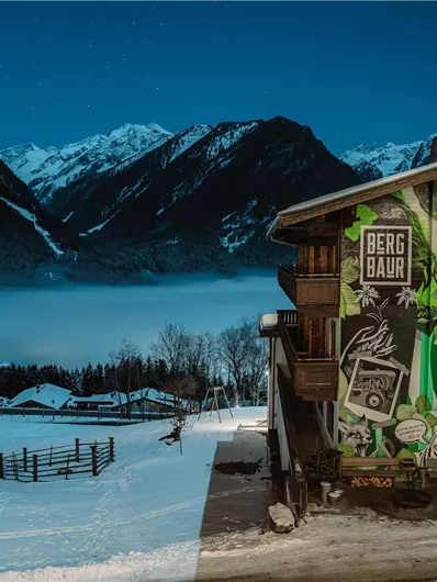 A modern old wooden house with a colorful wall design. In the background, snow-covered mountains and a foggy valley can be seen.