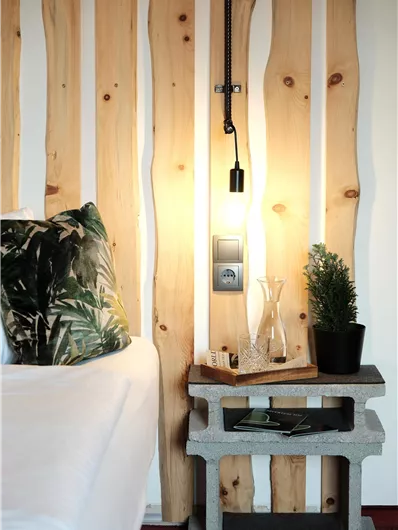 A modern bedroom with wooden paneling and a stylish bedside lamp. The concrete nightstand has decorative plants and a water container.