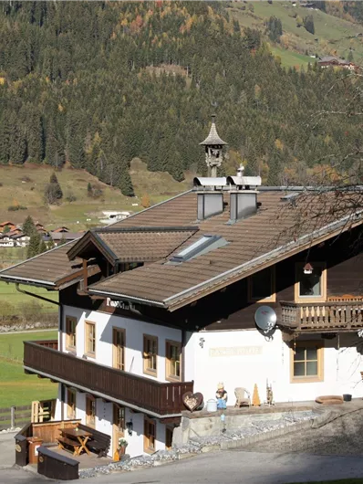 Ein charmantes Berghaus mit einem großen Balkon und traditioneller Architektur. Die umliegende Landschaft ist grün und hügelig, ideal für die Natur.