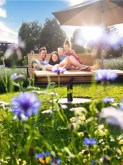 Een groep van vier mensen zit ontspannen op een ligstoel in de open lucht. Om hen heen bloeien mooie, kleurrijke bloemen en op de achtergrond zijn zachte heuvels te zien.