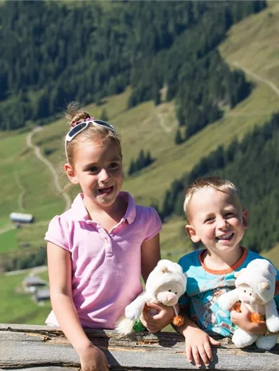 Twee kinderen staan aan een houten reling en houden knuffeldieren vast. Op de achtergrond zie je een mooie, groene berglandschap.