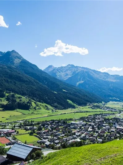 Een pittoresk landschap met groene weilanden en majestueuze bergen. In het dal liggen kleine dorpen onder een heldere blauwe lucht.