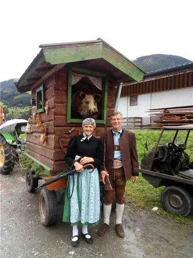 Een traditioneel paar in kostuums staat voor een klein houten huis op wielen. Daarachter kijkt een beer uit het raam.